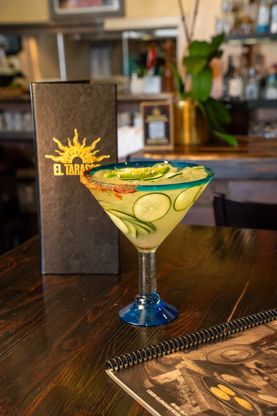 A cucumber margarita with a chile-salted rim in a blue-stemmed glass at the bar