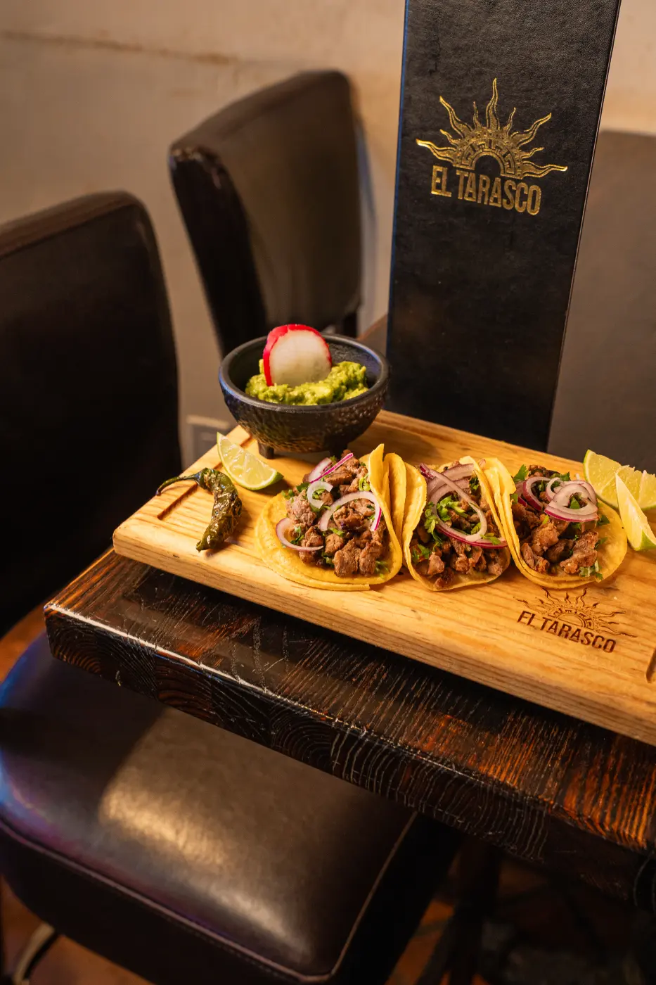 Three street tacos with cilantro and pickled onion on a branded board, served with guacamole, lime and a grilled chile