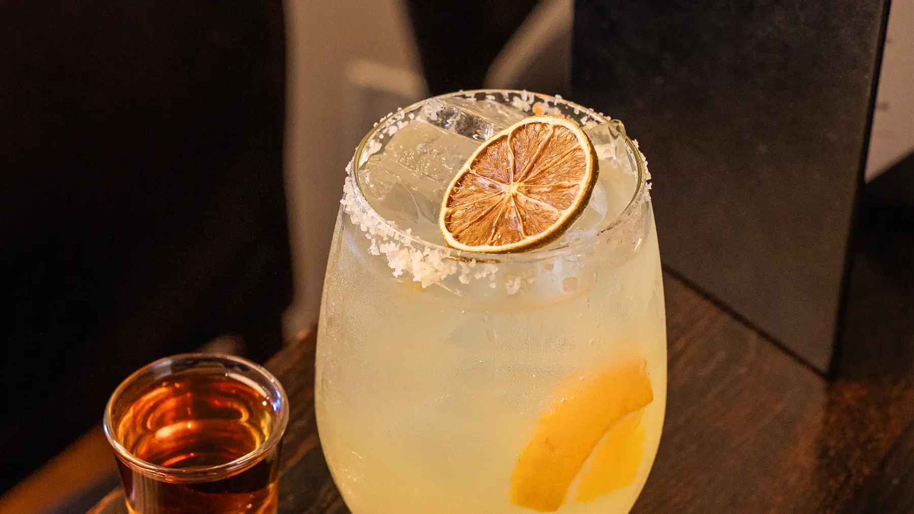 A salt-rimmed margarita with a dried lime wheel, poured alongside a shot of tequila
