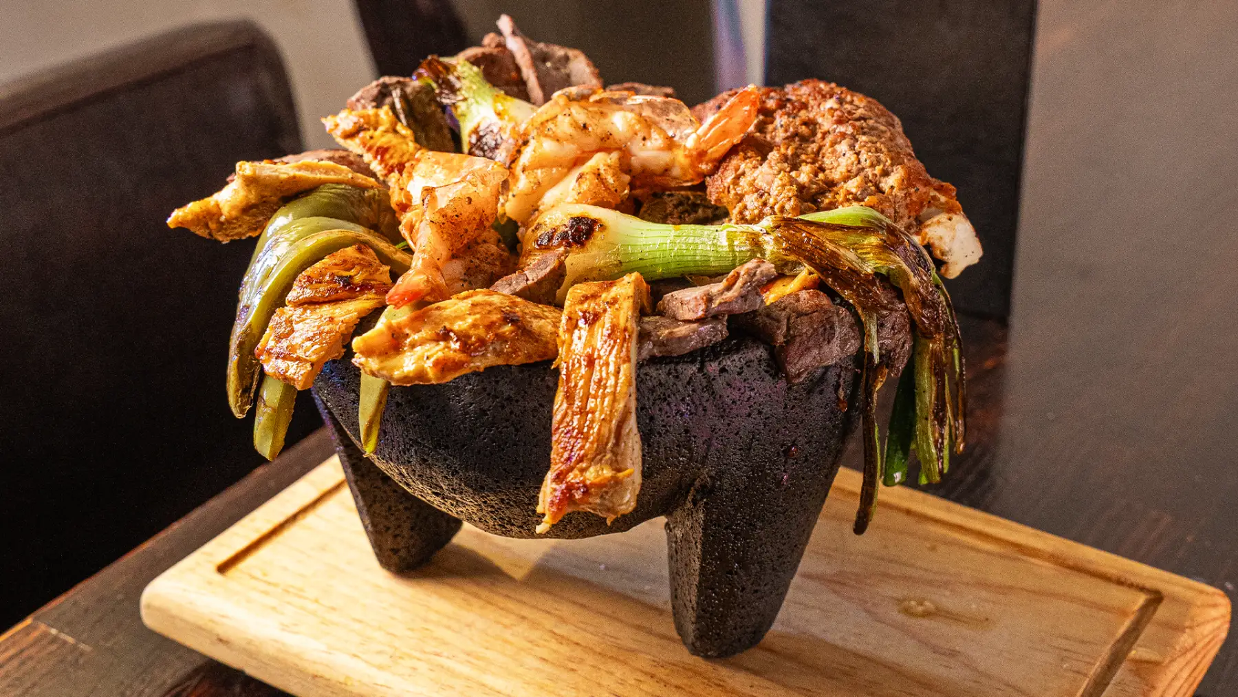 A molcajete piled with grilled shrimp, chicken, steak, cactus and spring onions on a branded El Tarasco board