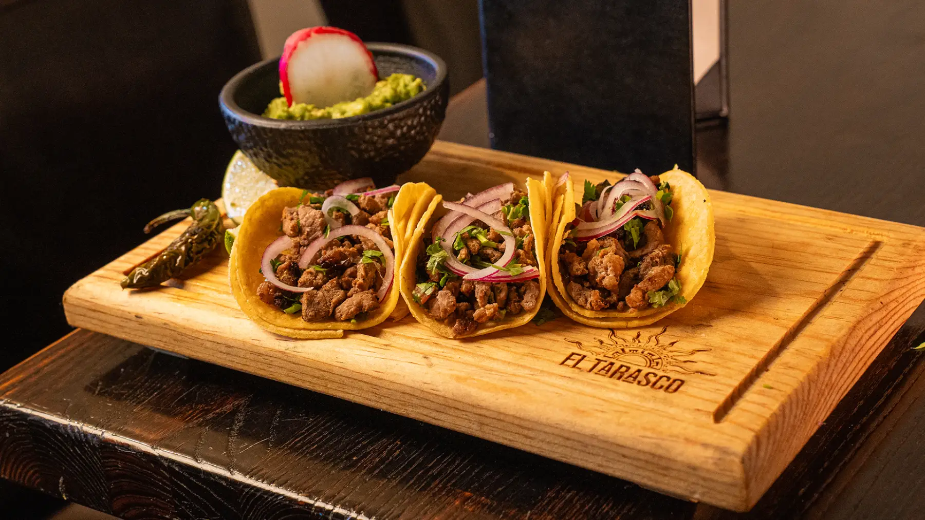 Three street tacos with pickled onion and cilantro on a branded El Tarasco board, served with guacamole and a grilled chile